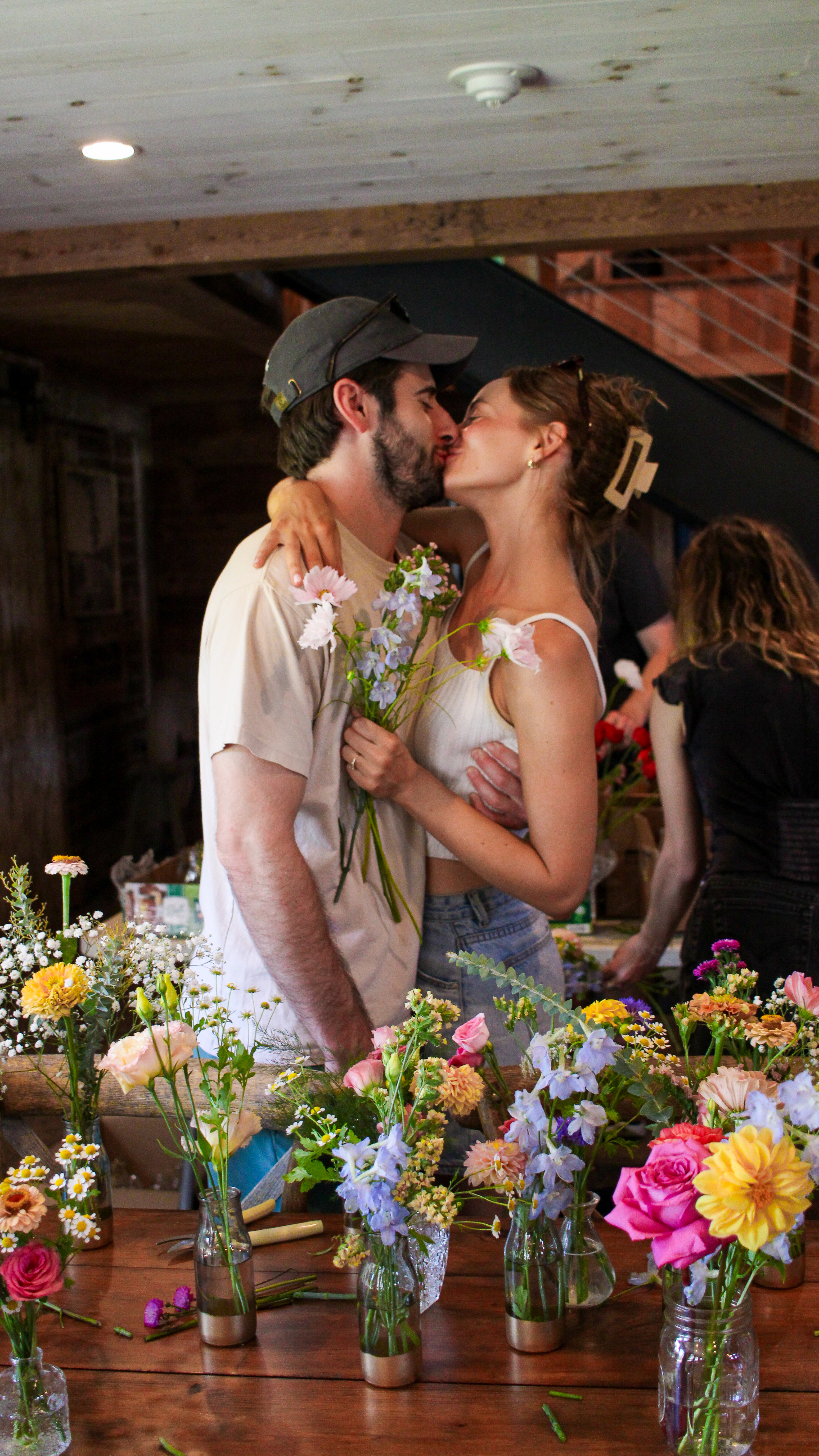 A couple kissing behind a table of freshly arranged wildflower bouquets