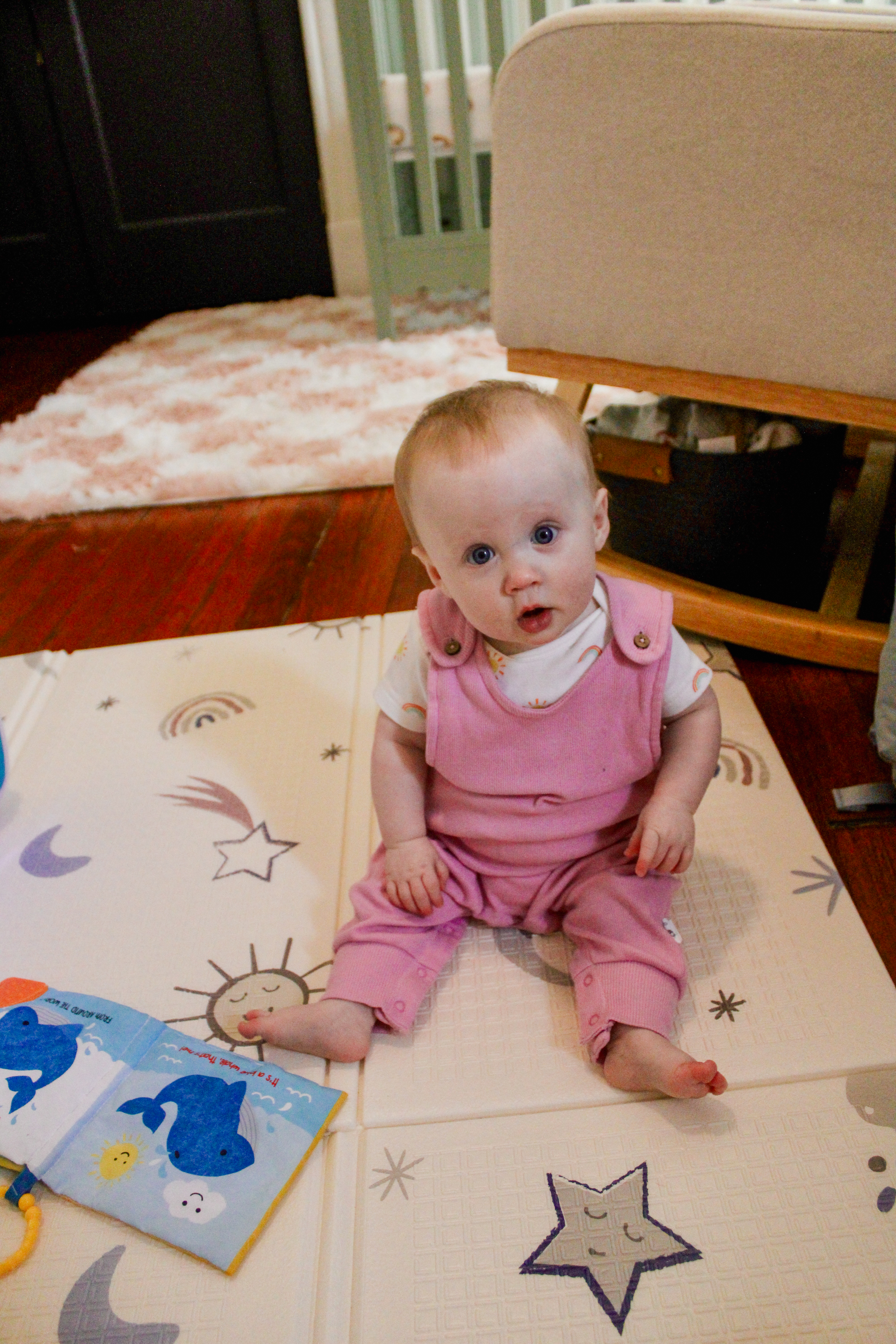 A baby in pink overalls sitting on a patterned play mat