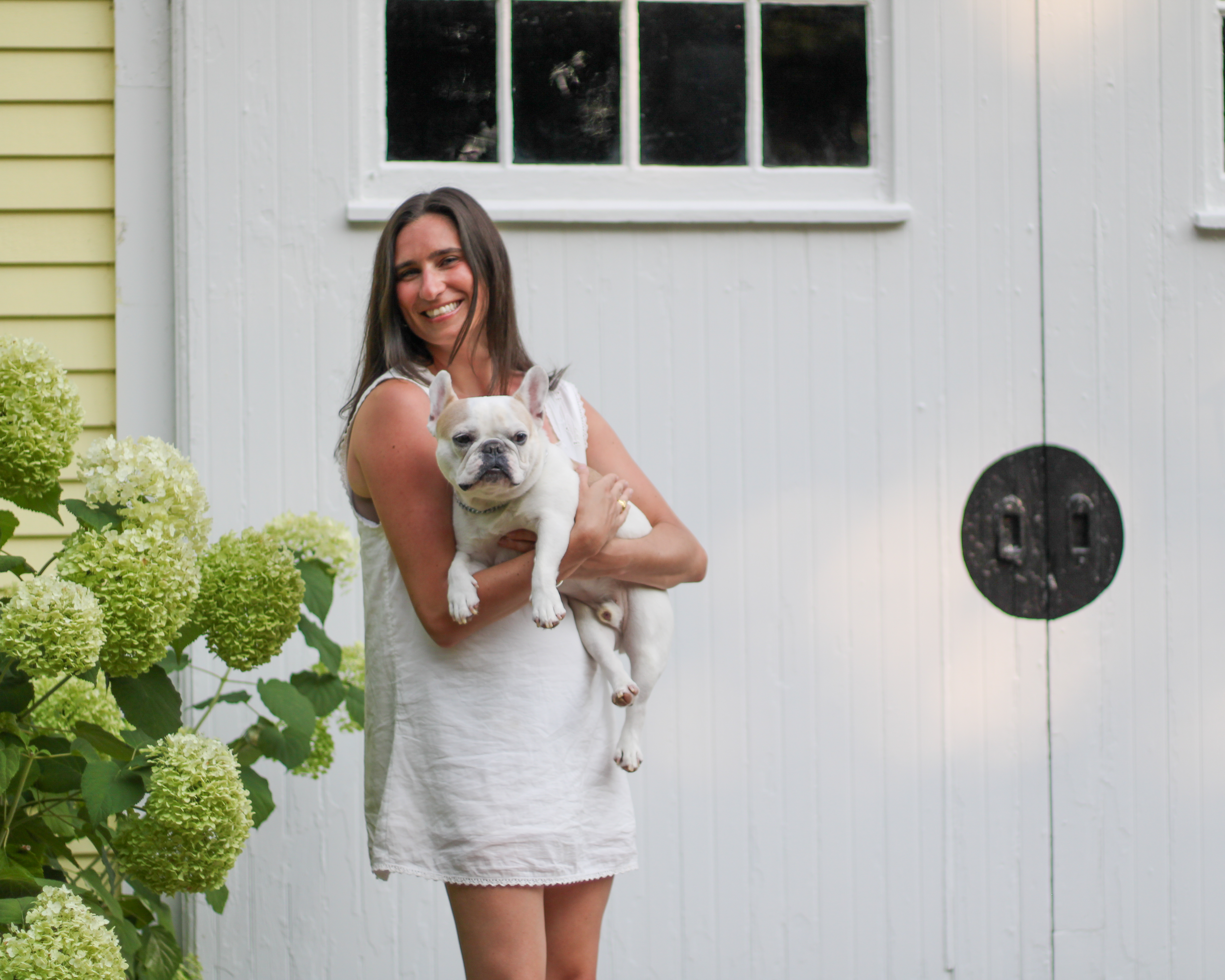 Sarah standing and holding her French bulldog by a white barn door