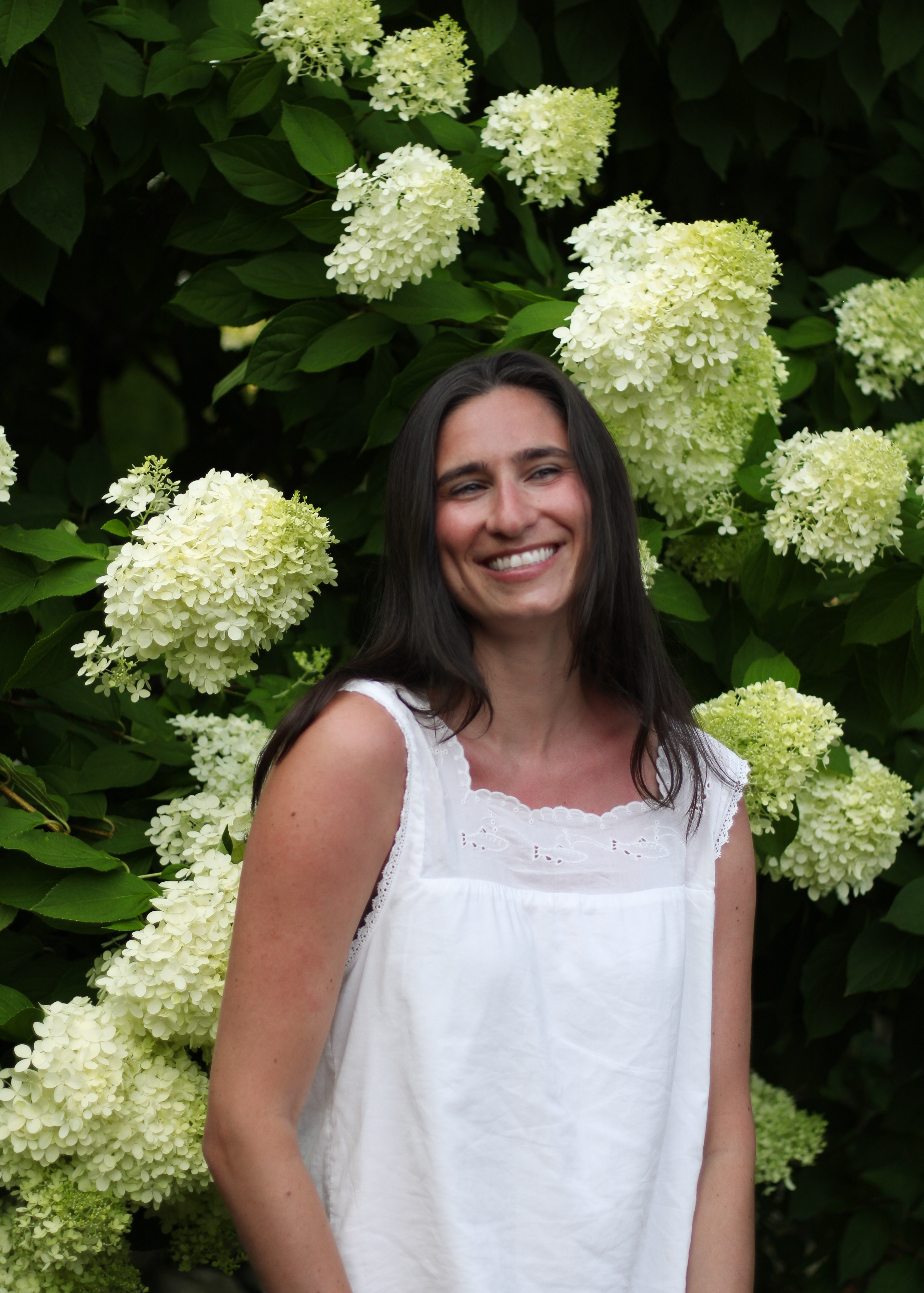Sarah standing among tall white hydrangea blooms in a white dress