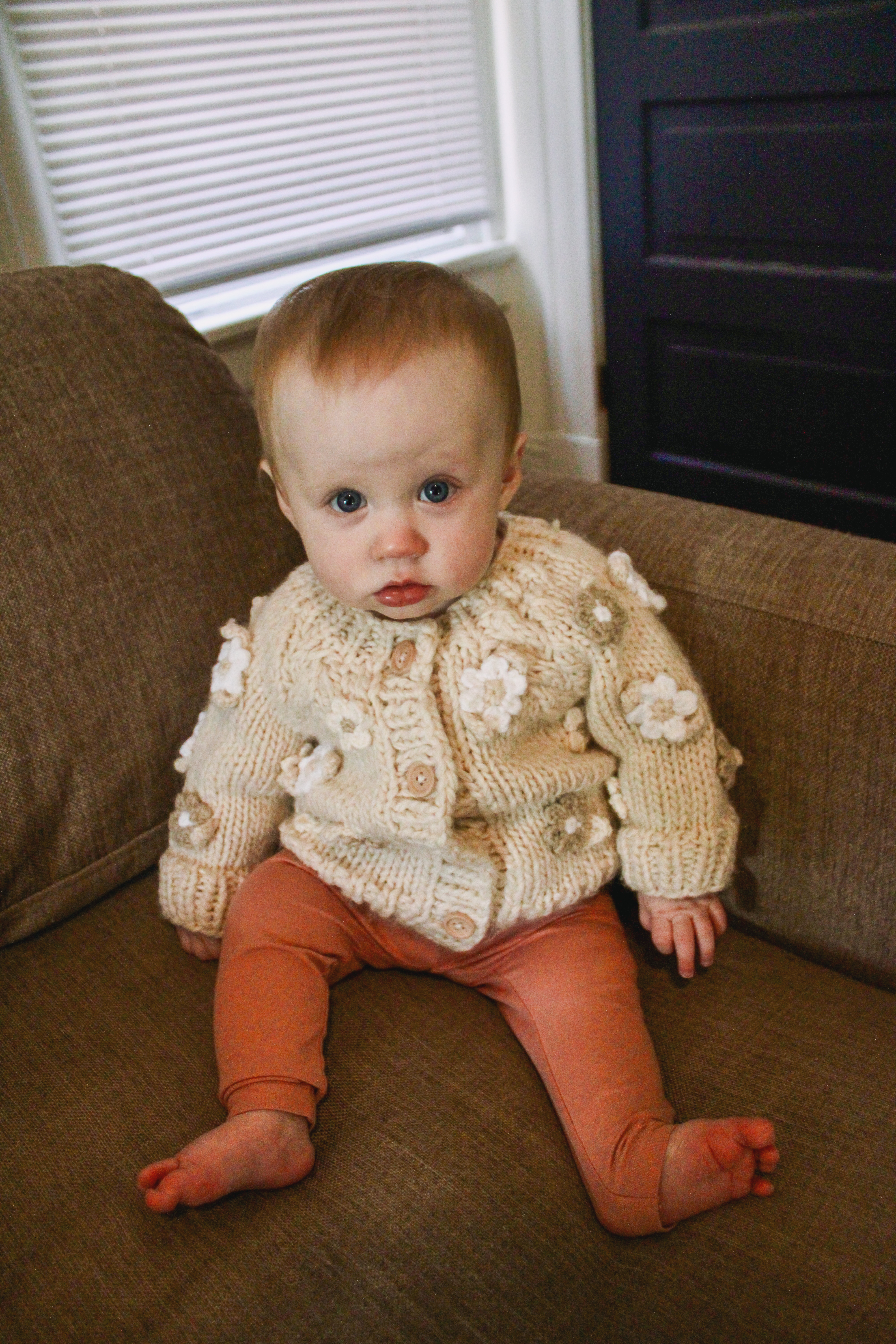 A baby in a cream knitted cardigan sitting on a brown couch