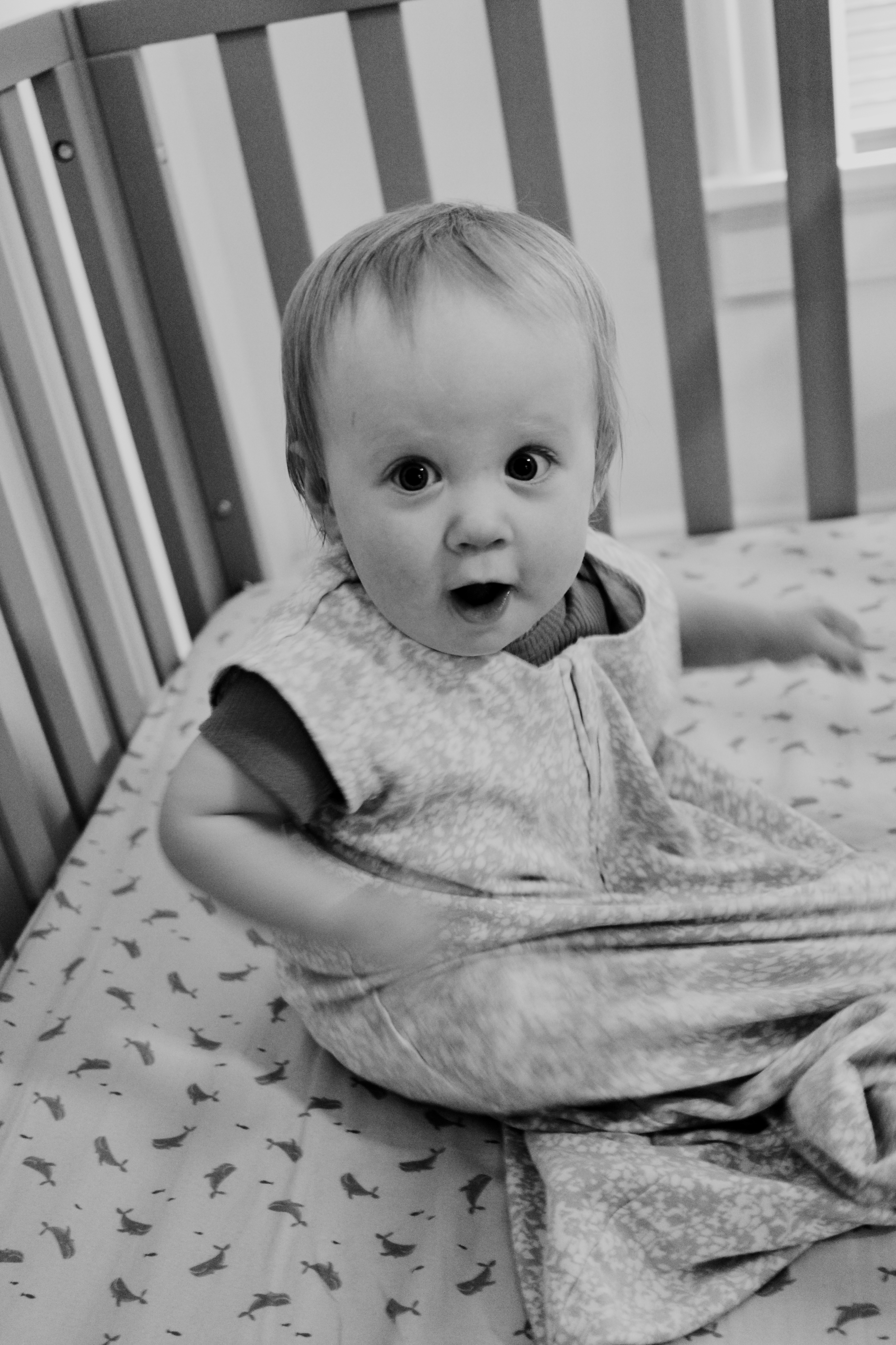 Black and white photo of a baby in a sleep sack sitting in a crib