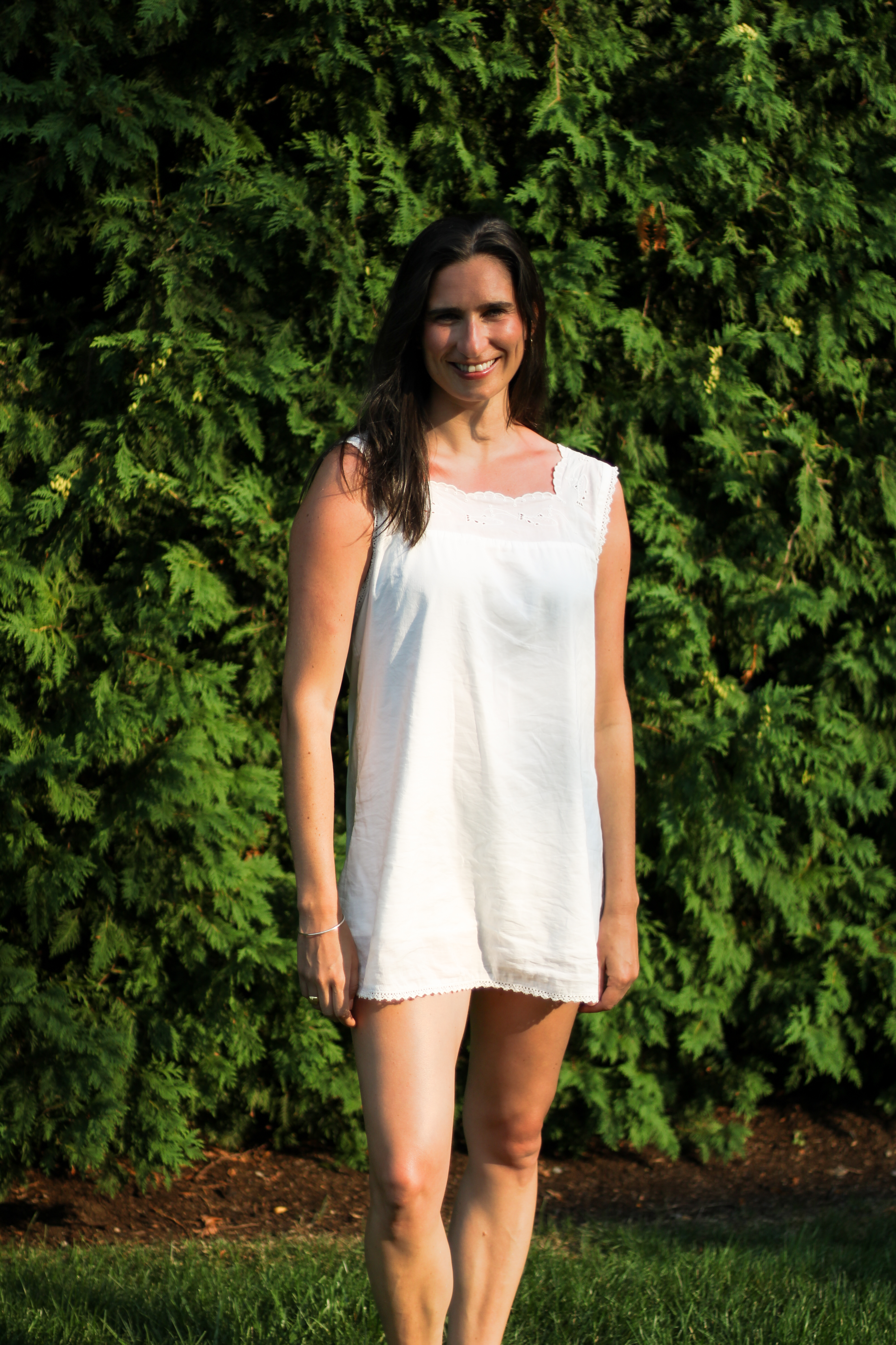 Full-length portrait of Sarah in a white dress in front of a tall green hedge