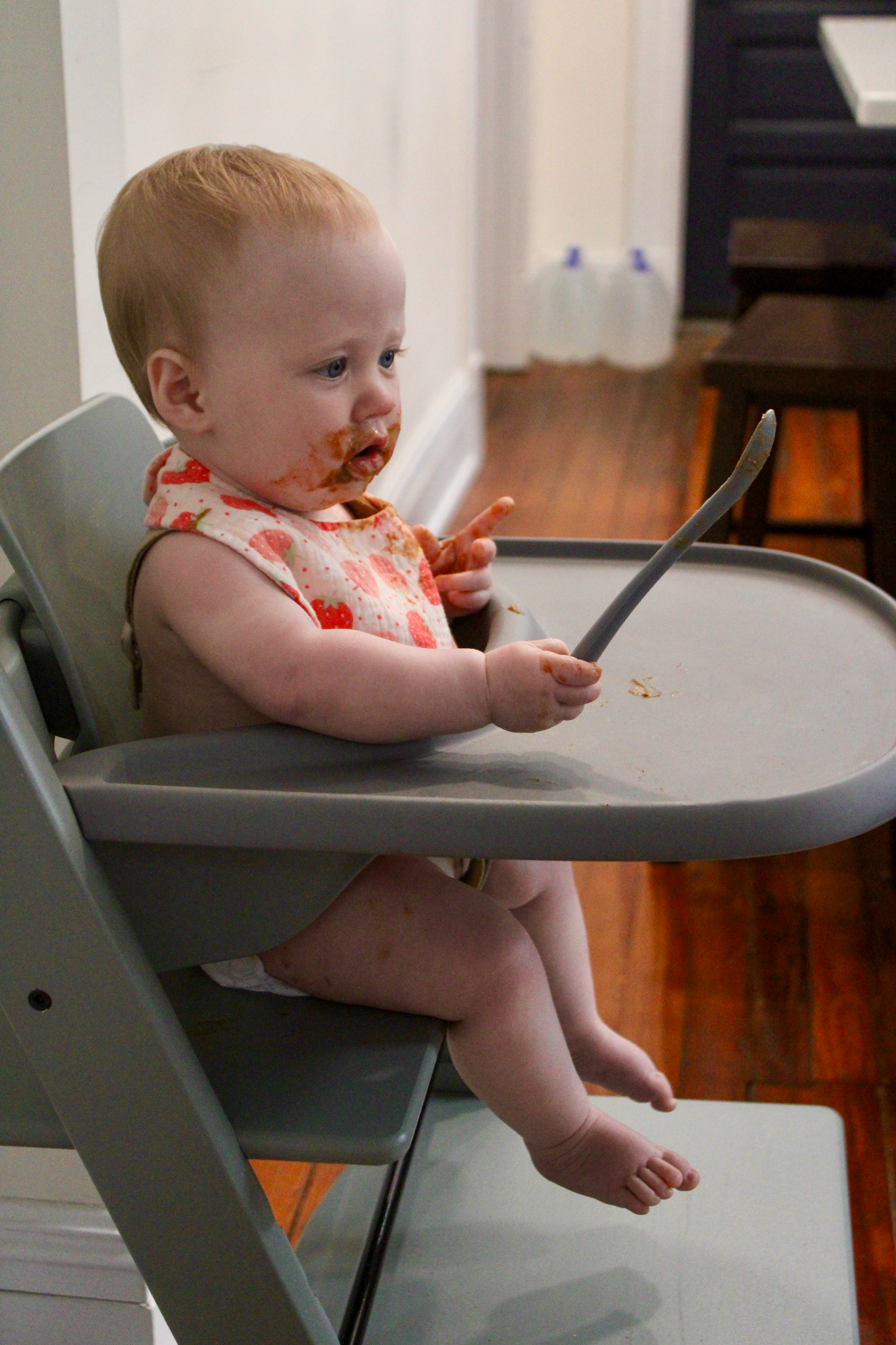 A baby holding a spoon in a high chair with food on her face