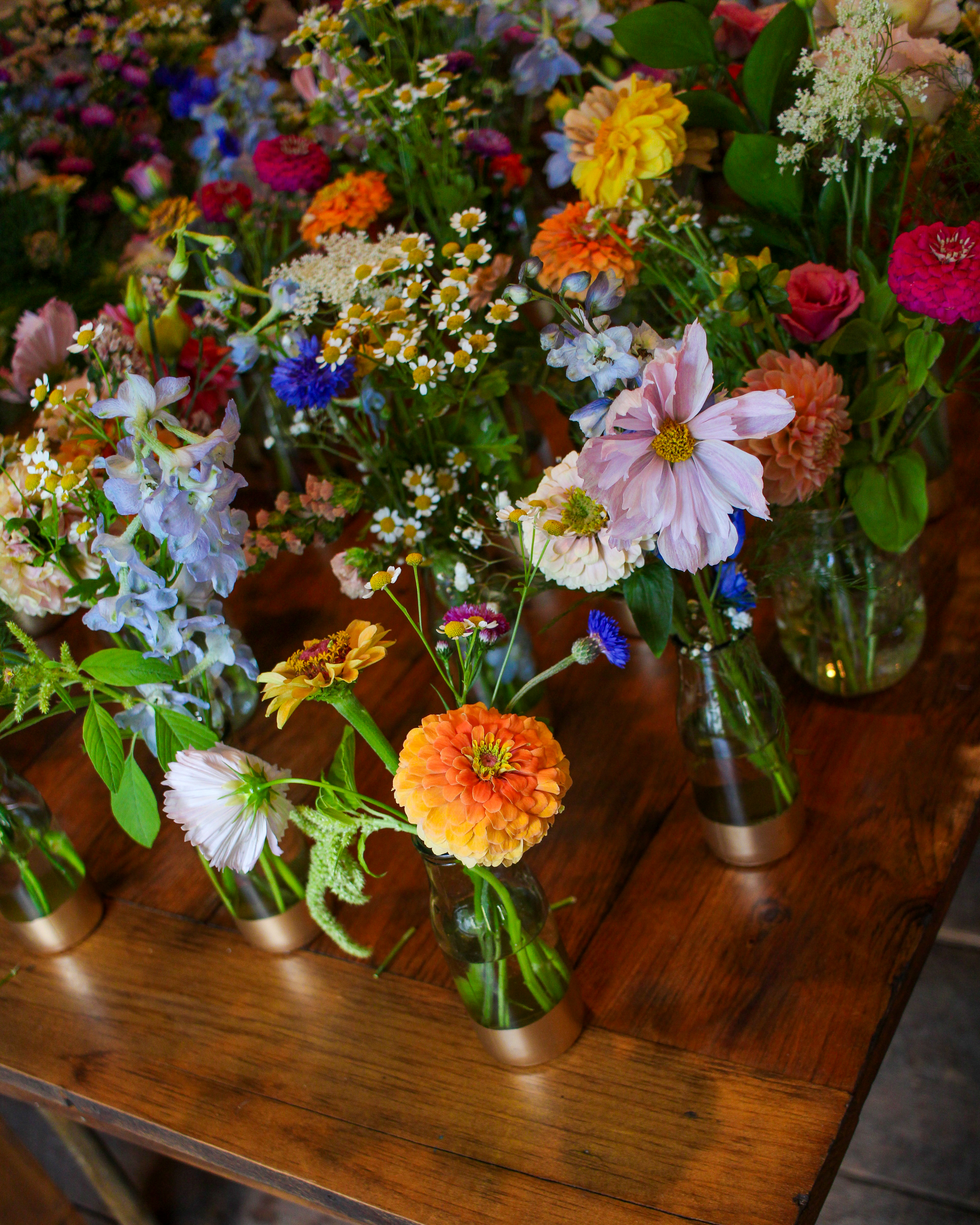 A long wooden farm table lined with wildflower bouquets in glass vases inside a barn