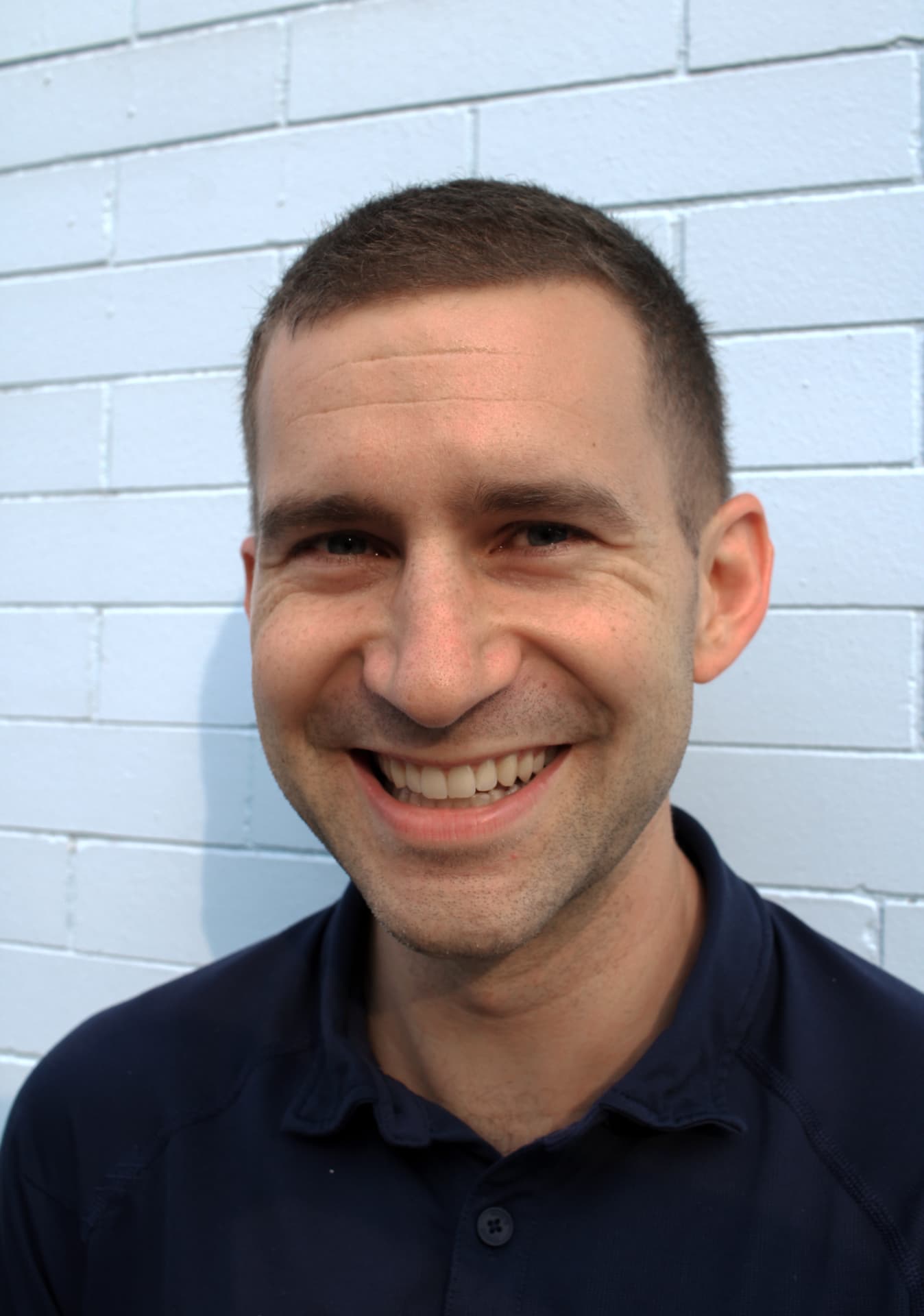 Smiling headshot in a navy polo in front of a white painted brick wall