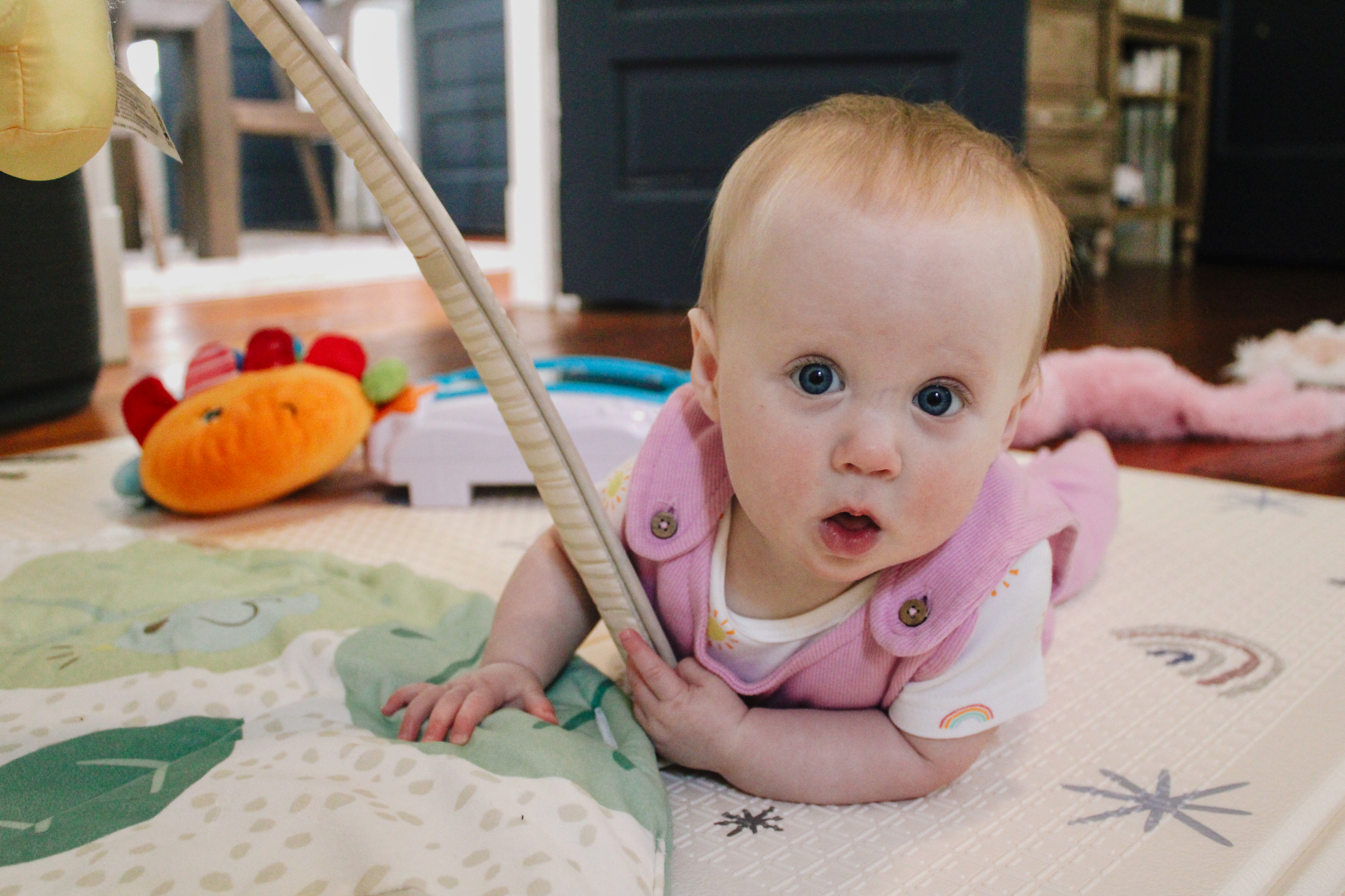 A baby on her tummy under a play gym, looking up curiously